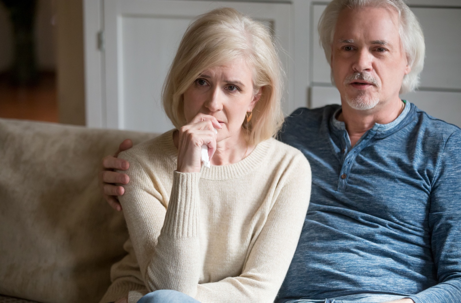 An older couple look concerned as they sit on a couch and watch a movie.
