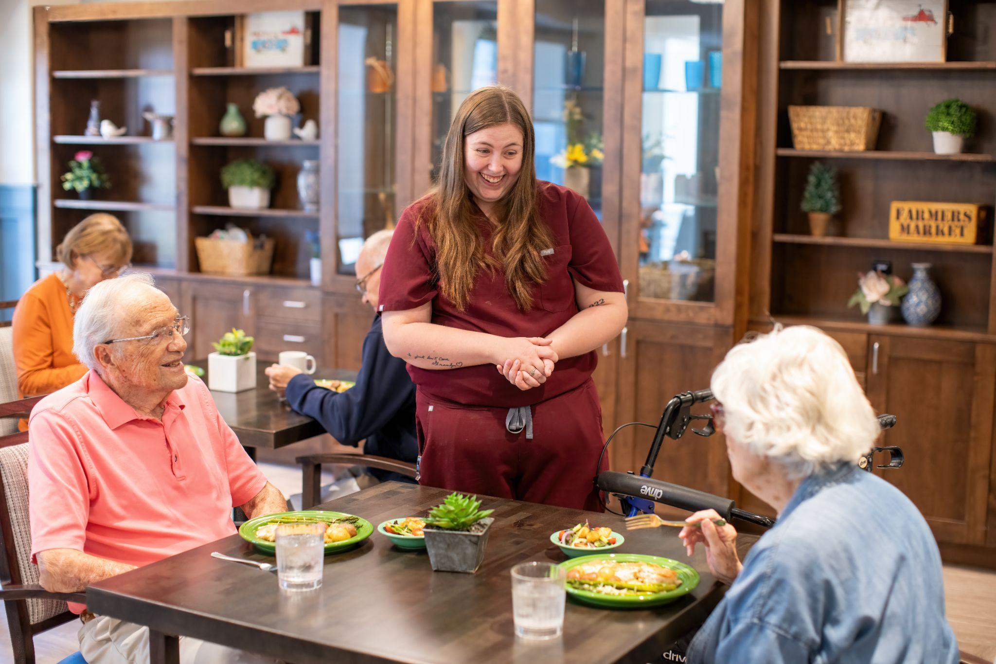 A smiling caregiver at High Plains Crossing chats with senior residents as they eat a meal.
