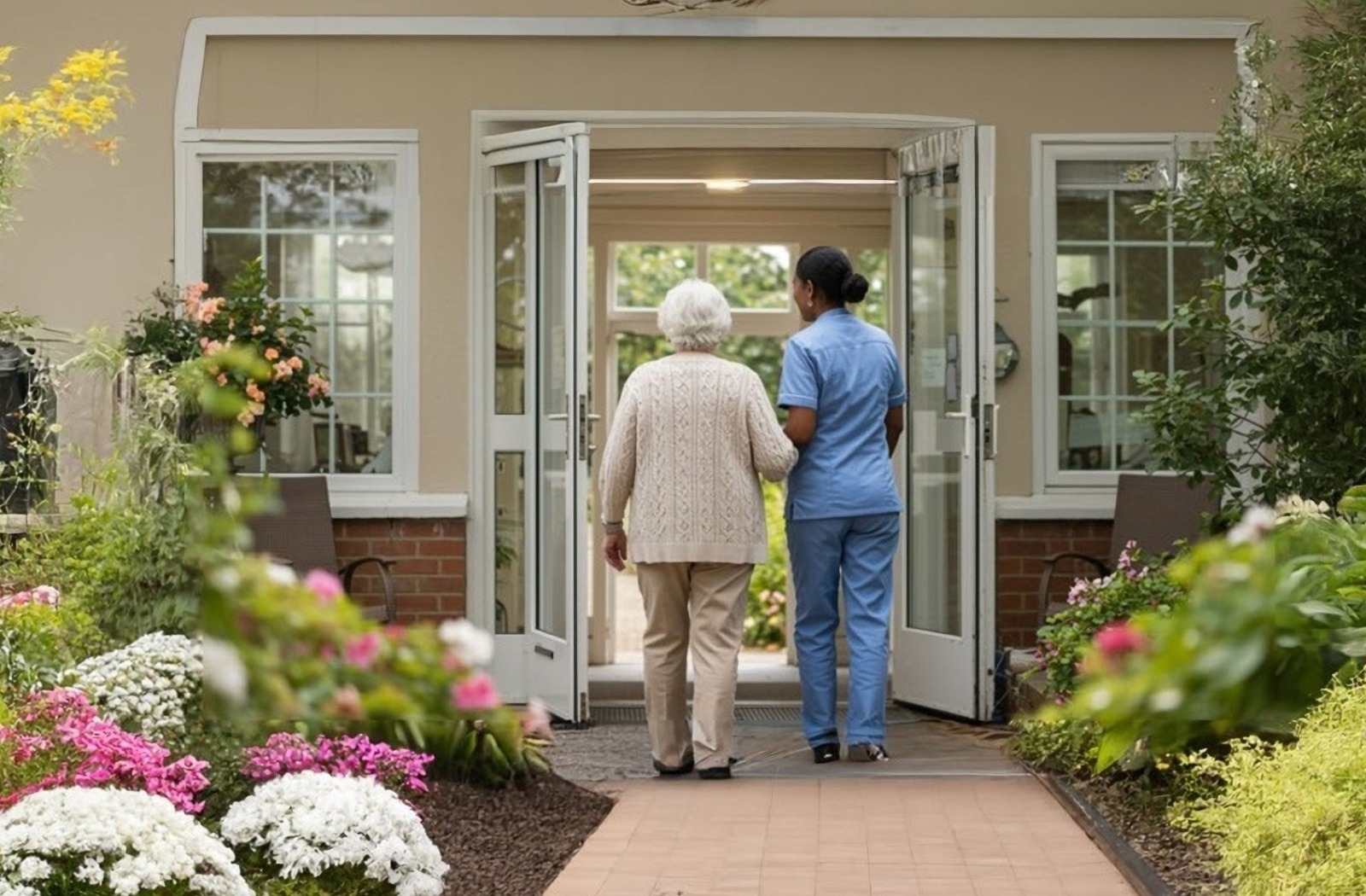 Elderly woman supported by caregiver in blue scrubs walking through open doors of a sunny, flower-filled care home entrance.
