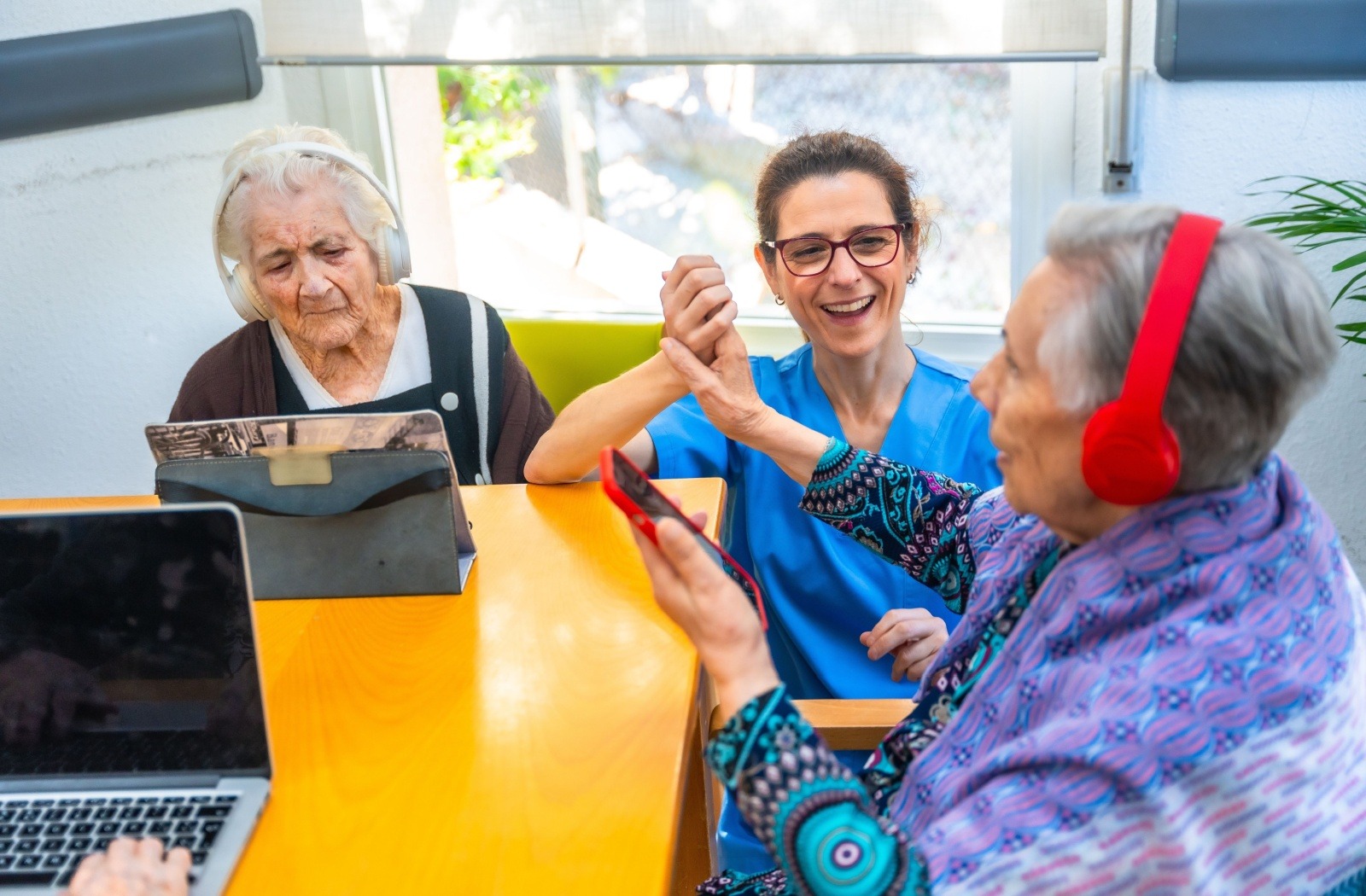 Caregiver and two elderly women share a joyful high-five at a bright table, wearing headphones in a social activity room