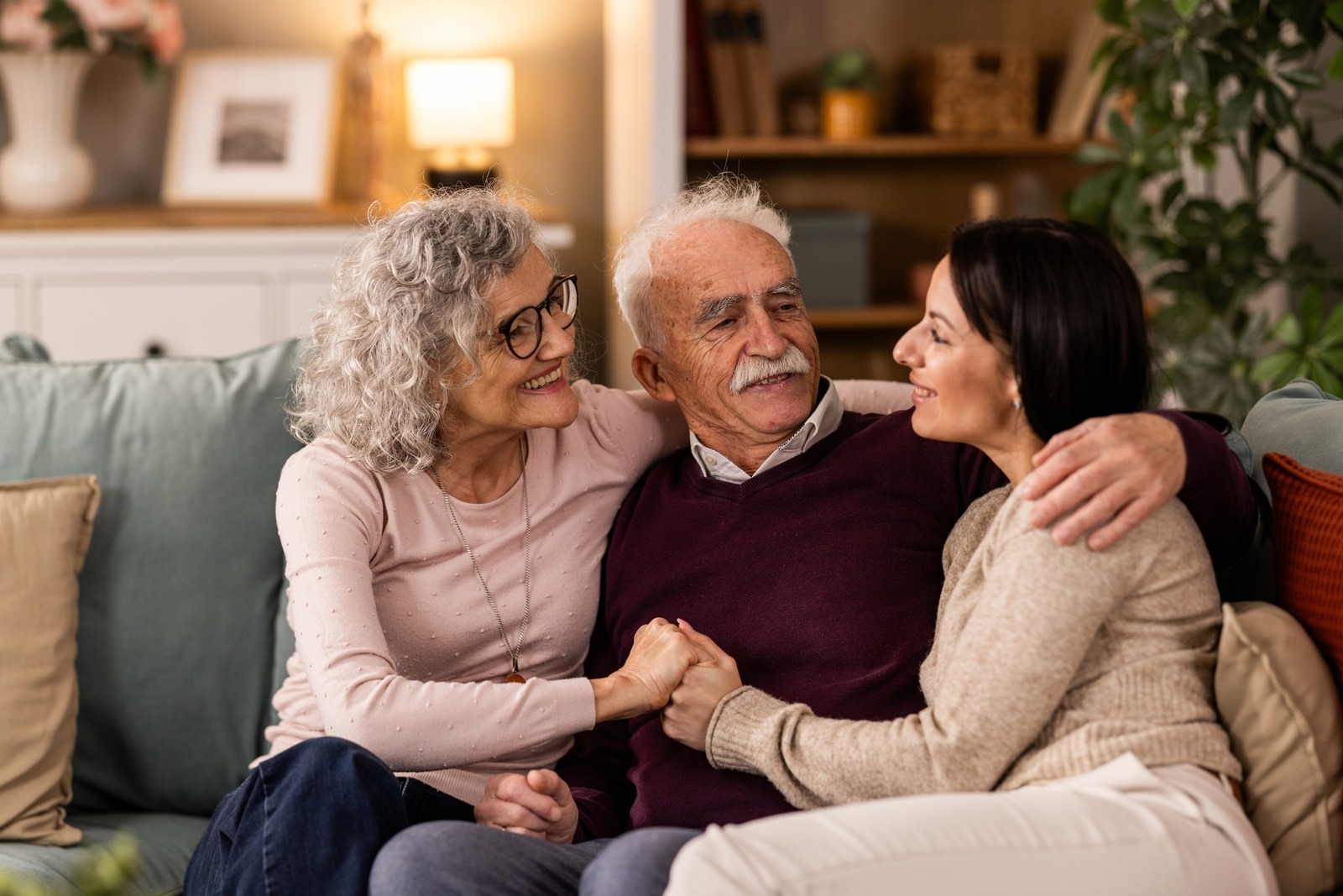 Smiling daughter hugging with her senior parents while she is visiting them, they are sitting on the couch in the living room.