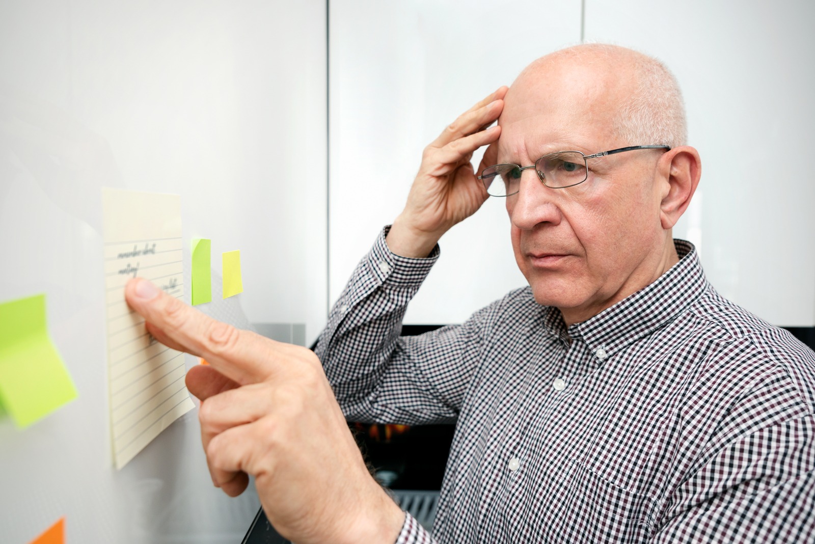 Older man with glasses points to lime sticky notes on a glass board, thinking deeply.